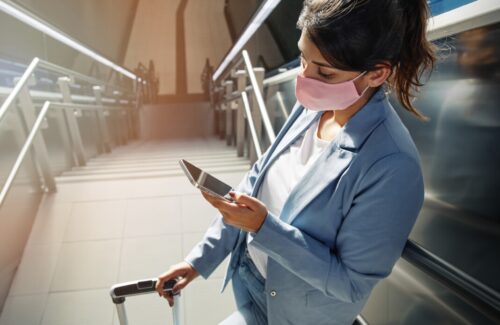 high angle of woman with medical mask and luggage using smartphone at the airport during pandemic